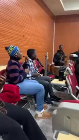 The video shows a small group of people rehearsing in what appears to be a church setting. The room has wooden walls, with a cross displayed on the wall behind the keyboardist. There are seven people visible, each engaged in their respective roles. Two singers are seated, sharing a microphone. A musician playing a bass guitar is behind them. A man is playing drums in the background. Seated next to the singers, a person is holding a microphone, possibly singing. The final person visible is playing a keyboard and wearing a mask. The furniture includes red and white chairs and tables supporting music equipment. The atmosphere seems relaxed and collaborative.