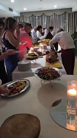 A group of people are gathered around a buffet table in a room with dim lighting and draped curtains. In the foreground, a woman in a black and white floral top and glasses is serving herself food from a silver platter. To her left, another person is also serving themselves from a platter. The buffet table is laden with various dishes, including a large roasted pig, bowls of colorful food, and platters of appetizers. Several staff members in formal attire are assisting guests and replenishing the food. Candles are placed on the table, creating a warm ambiance. The environment appears to be a restaurant or event hall.