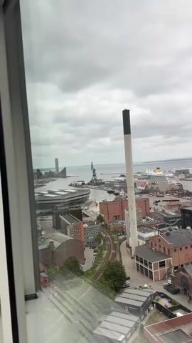 A vertical video shows a panoramic view of a city skyline from a high vantage point, possibly a window. The view encompasses a body of water in the distance, with a harbor featuring cranes, ships, and buildings. Closer to the foreground, there are numerous buildings of varying heights and architectural styles, including a tall, slender smokestack, a modern skyscraper with a distinctive white facade, and several brick buildings. Some buildings appear to be under construction, as indicated by a prominent crane. The sky is overcast with clouds. The video pans from left to right, revealing more of the city and the waterfront.