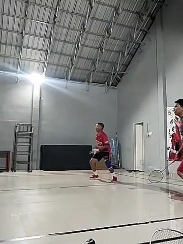 Two men are playing badminton indoors. The man on the left is wearing a red shirt and black shorts, and is in motion, swinging a badminton racket. The man on the right is wearing a blue and red patterned shirt and red shorts, and is also in motion, holding a badminton racket. The court has a grey floor with white lines. The ceiling is made of metal panels and has industrial lighting. There are metal shelves and boxes in the background.