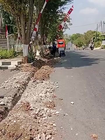 This is a video captured from a slightly elevated, moving perspective, looking down a paved road. On the left side of the road, a trench has been dug along the edge, with piles of dirt and rocks beside it. A person, wearing a dark shirt and pants, is actively working in the trench with what appears to be a shovel. Nearby, there's an orange truck, a generator, and other equipment. Several trees line the left side of the road, decorated with red and white striped flags. The road itself shows some wear and tear. In the background, more trees and buildings are visible, and several motorcycles are traveling down the road. The overall environment appears to be a semi-urban or rural setting on a sunny day.