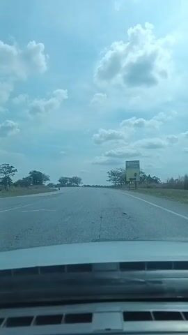 The video shows a car driving on an asphalt road on a partly cloudy day. The road is mostly straight with visible cracks and patches. There are sparse trees on either side of the road. A billboard is visible on the right side of the road. Another car is visible in the distance on the road ahead. The dashboard of the car recording the video is visible.