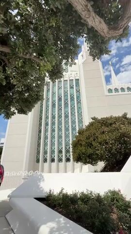 A low-angle shot pans upwards to reveal the exterior of a white, ornate temple-like building with tall, pointed spires. The building features large, vertical windows with a patterned green tint and intricate white architectural details. Lush green trees and shrubs surround the base of the structure, and the sky above is a vibrant blue with scattered white clouds. The camera moves from a close-up of the building's facade to a wider view of its structure against the sky.