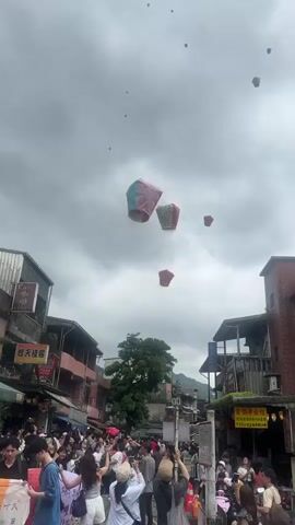 A street crowded with people looking up at several sky lanterns floating in the sky is shown in this video. The sky is overcast with gray clouds. The buildings are varied in style, with some featuring signs in a non-English language. The lanterns are different colors, including red and teal, and are dispersed across the sky. The crowd appears dense, with many individuals holding cameras or phones, presumably to capture the sight. A large green tree partially obscures some buildings in the middle ground. The overall atmosphere suggests a festive or celebratory event.