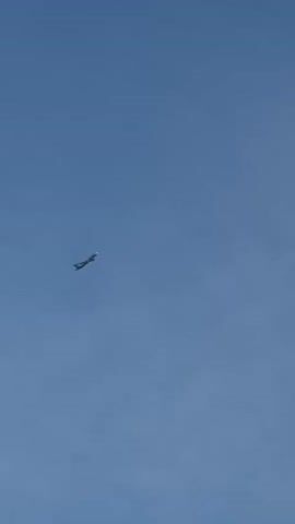 The video shows a single airplane flying through a clear, bright blue sky. The airplane is a light color, possibly white or silver, with dark accents, and it appears to be climbing. There are no clouds visible, suggesting a clear weather day. The setting is an open sky, with no other objects or land features visible. The time of day appears to be daytime due to the brightness of the sky. No people are visible in the video. The overall mood is serene and unremarkable, focusing on the solitary flight of the aircraft. No text is visible in the video. The location context provided is "Дольче Вита Кп, Russia," which indicates a geographical area but offers no visual cues within the video itself to confirm or specify this location.