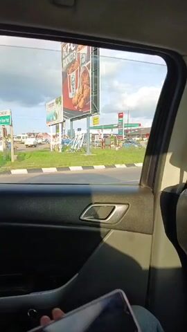 From inside a vehicle, the camera shows the outside world through a car window.  The scene includes a KFC billboard advertising buckets of chicken, a PUMA gas station, Pizza Hut and KFC stores, cars, and other signs. The sky is partly cloudy. The car door interior is visible in the foreground. The vehicle is moving.