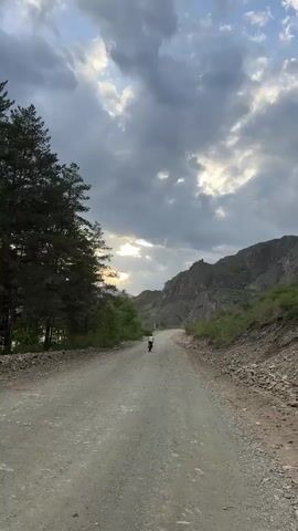 A single person is cycling away from the viewer on a dirt road. The road is surrounded by mountains and trees. The sky is cloudy, but there is a patch of light shining through. The person is wearing light-colored clothing and is moving away from the camera. The surrounding environment includes rocky mountains and greenery, suggesting a remote or mountainous area. The overall tone is tranquil, capturing the solitary activity within a vast landscape.