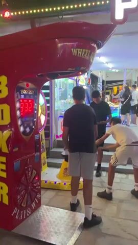 A group of young males are gathered around a large, red arcade game machine labeled "BOXER." The machine features a digital display showing "332" and has a circular spinning wheel with various prizes. One male, wearing a black t-shirt and grey shorts, stands facing the machine. To his right, another male in a black t-shirt and shorts holds a mallet, positioned as if to strike the machine. A third male, in a white t-shirt and shorts with a black design, also holds a mallet. They appear to be waiting for their turn or observing the game. The environment appears to be an outdoor amusement area with string lights overhead.