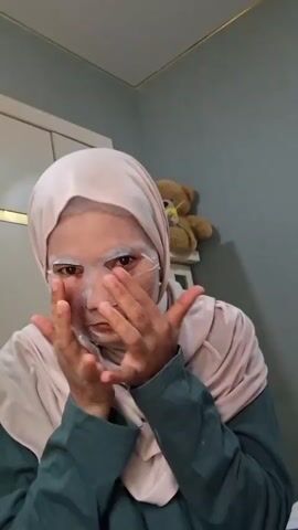 A person wearing a hijab and a green long-sleeved shirt applies a SKINTIFIC Niacinamide Brightening Daily Mask to their face. The person adjusts the mask on their face, smoothing it out with their hands. They then hold up the pink box of face masks for the camera, smiling slightly. A white cabinet and a teddy bear on a shelf are visible in the background. The room is painted a muted grey-blue color.