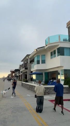 The video captures a beach scene on a cloudy day. Several houses line a path, with a person walking a dog on the sand side and two individuals strolling along the path. The beach features people in the distance, and a person throwing a frisbee near the water's edge. The sky is overcast, with hints of sunlight breaking through near the horizon.