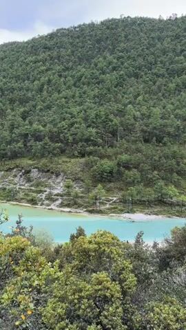 This is a vertical video of a scenic landscape with a turquoise lake and a forested mountain. In the foreground, there are trees with green leaves obscuring part of the view. The middle ground shows a body of turquoise water with a sandy bank. Along the bank, there are walking paths and a few small trees. In the distance, a lush green mountain covered in dense forest rises steeply. The sky above is cloudy. Towards the right side of the lake, there is a bridge and a gathering of people, some of whom appear to be walking or standing near the water's edge. The video pans slightly to the right, revealing more of the lake and the mountainous terrain.