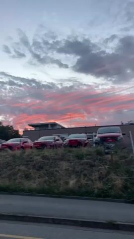 A low-angle video shows a row of red cars parked on a grassy incline with a building in the background. The sky above is filled with dramatic clouds tinged with pink and orange from a sunset. A tall lamppost stands to the left of the frame, and trees are visible behind the cars. The video pans slightly to the right, revealing more of the parked cars and the building. The overall impression is of a serene evening scene with a vibrant sky.