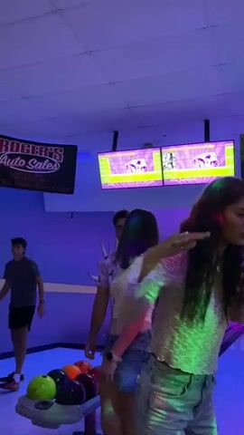 The video shows a group of people in a dimly lit bowling alley. The environment is characterized by purple and blue neon lighting and several screens displaying what appear to be scores or game information. In the foreground, a young woman with long dark hair and wearing a sparkly top and denim shorts is visible, facing towards the right of the frame and appearing to be speaking or reacting. Behind her, another young woman in a white crop top and denim shorts is seen from the back, facing the bowling lanes. Further back, other individuals are scattered around, some near the bowling lanes. A bowling ball and pins are visible in the lower left corner of the frame. The overall atmosphere suggests a social gathering or a night out at a bowling alley.