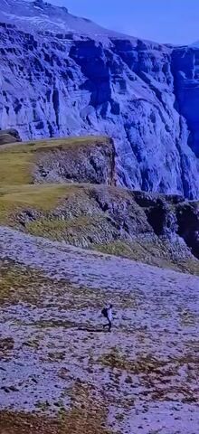A person in a white top and dark pants runs down a steep, dry grassy slope in front of a large, purple cliff face. The sun casts a long shadow of the person onto the ground.