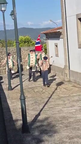 The video shows a group of people parading down a cobblestone street. They appear to be marching band members, several wearing traditional costumes with large drums strapped to their chests. Some of the marchers are young, perhaps scouts. One person carries a tall flag with a unique design consisting of red, white, and blue elements. The street is lined with old-fashioned lampposts and bordered by a stone wall on one side and a white building on the other. In the background, rolling green hills can be seen under a clear blue sky. The shadows from the lampposts stretch across the cobblestones, indicating bright sunlight.