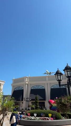 A wide-angle, vertical video captures an airplane descending against a clear blue sky. The plane is white with visible markings, including a blue and red symbol on its tail. Its landing gear is extended, indicating it is preparing for landing. Below the airplane, a modern building with classical architectural elements is visible. The building has large arched windows and ornate columns. In front of the building, there is a landscaped area with plants and flowers, and a circular planter featuring a large, colorful heart-shaped sculpture made of rainbow-colored panels. Several people are casually walking and sitting in this area, enjoying the outdoor environment. The scene suggests a shopping or entertainment district located near an airport. The sun is shining brightly, casting clear shadows and illuminating the scene.