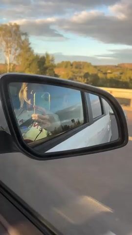 The video shows a view out of a car window, focusing on the side mirror. The side mirror reflects the passenger in the car, who is holding a phone and taking a selfie. The passenger is wearing a light-colored shirt with some writing on it. The car is moving, as indicated by the blurred background and the road markings visible outside the window. The background seen in the mirror and outside the window consists of trees, fields, and a cloudy sky, suggesting it is daytime. The overall impression is a person capturing a moment during a car journey.