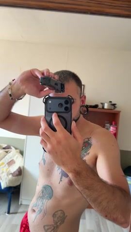 This media file features a young man in a dimly lit room, possibly a bathroom or bedroom, as indicated by the mirror and furniture in the background. He is shirtless, revealing several tattoos on his torso, including a jellyfish and a skeletal figure. He is holding a mobile phone with a prominent camera array in his right hand, using it to take a selfie in the mirror. His left hand holds a hair clipper, which he is using to shave the top of his head.

The man appears to be in the process of giving himself a haircut. The lighting suggests it might be daytime, but the indoor setting makes it difficult to determine. He has ear gauges and is wearing red shorts. The background shows a closet with some items on shelves, a bed with a quilt, and a trash can. The overall impression is a personal moment of grooming. There is no visible text in the media.