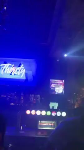The video shows a crowded nightclub with a bar and a dance floor. The club has a DJ screen, chandeliers, and dynamic lighting. There are people dressed in casual clothes, milling around and talking. The camera moves around a bit, showing different areas of the club. The video appears to have been taken at night.