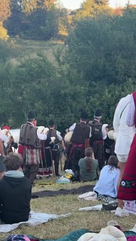 A group of people in traditional Bulgarian folk costumes are gathered outdoors, seemingly in a grassy field with trees in the background. The men are wearing dark hats, vests over white shirts, and patterned skirts or kilts, some carrying what appear to be bagpipes. The women are also in colorful traditional attire, with one woman in the foreground wearing a red vest over a white shirt and a long, embroidered skirt. Several spectators are seated on the ground watching the performers. The overall scene suggests a cultural performance or celebration.