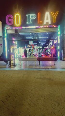 A vertical shot depicts a building exterior at night. The sign "GO PLAY" is illuminated vertically with neon lights. The building's interior is visible through large glass panels, revealing arcade games and other entertainment equipment. A person in dark clothing stands inside, seemingly observing the scene. A wooden bench sits outside the building along a brick-paved sidewalk.