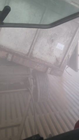The video shows a view from inside the cab of a vehicle, possibly a tractor or other agricultural machine. The view through the windshield is heavily obscured by dust or fog. We see the back of a truck or trailer bed, partially visible through the haze, with a sticker labeled "51034". The vehicle appears to be driving over a metal grid or grate. Inside the cab, we see the control panel, levers, and the interior structure of the cabin. The camera angle changes, showing the dashboard and the metal frame of the cab.