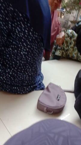 The video shows a close-up view of people sitting on a floor, with a small pink purse in the center of the frame. In the background, a person wearing a blue and white patterned dress and a dark blue headscarf is visible. To the left, another person in a red dress and a light green headscarf is seen sitting on a bench. To the far left, two people wearing light purple pants and a white shirt are also visible. The floor is tiled in a light beige color. The overall setting appears to be a public waiting area or a communal space.