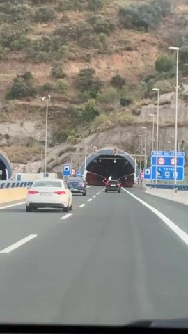 A POV shot from inside a car shows a highway approaching a large tunnel entrance carved into a hillside. The tunnel mouth is illuminated, with traffic lights visible above it. Several cars are driving on the highway, including a white sedan in the left lane and a dark-colored vehicle ahead in the right lane. Road signs with blue backgrounds and white text are posted on the right side of the road, indicating tunnel information and speed limits. The hillside is covered with sparse vegetation. The sky is overcast. The video is short and shows no distinct interactions between people or objects.