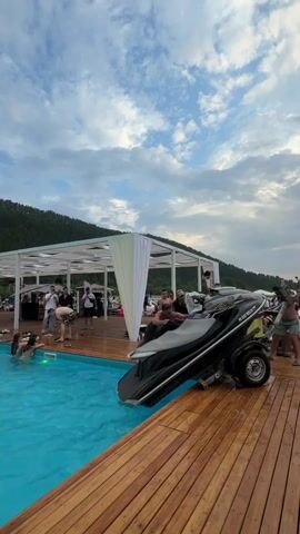 The media file depicts a scene at a waterfront establishment, likely a resort or event space, in Drokino, Russia. The primary subjects are a group of people and a jet ski.

The scene is set on a wooden deck adjacent to a swimming pool filled with clear turquoise water. In the background, a white, open-sided structure with a canopy provides shade and appears to be a gathering area or bar, with several people mingling underneath it. Lush green hills rise in the distance, suggesting a natural, possibly mountainous, landscape. The sky is filled with dramatic, cloudy formations, indicating it's likely late afternoon or early evening.

The main activity centers around a black jet ski, which is partially submerged in the swimming pool, resting on a trailer. Several people are gathered around the pool and the jet ski, some in the water and others on the deck. Their postures and the general atmosphere suggest a relaxed, social gathering, perhaps a party or a leisurely afternoon by the water. One woman is visible in the pool, appearing to interact with someone near the jet ski.

Notable details include the juxtaposition of the jet ski, typically used in open water, being placed in a swimming pool. This unusual placement might be part of an event or a unique recreational setup. The lighting suggests a relatively low sun, casting soft light and creating gentle shadows.

Visible text on the jet ski reads "FLIGHTMASTER" and what appears to be a registration number "RX 0207 TB 2A".
