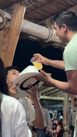 The video shows two young men in a social setting. One man is holding a white cowboy hat up to another man's face. The second man, wearing a light green shirt, is pouring a yellow beverage from a plastic cup into the hat. The man with the hat is drinking from it. Several other people stand nearby, observing the scene. The setting appears to be an outdoor event, possibly a bar or restaurant, with a wooden structure overhead. The lighting is dim, and there is an evident festive atmosphere with music in the background.