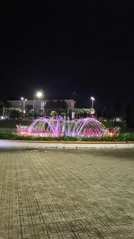 The video shows a fountain at night. The fountain is circular and has multiple jets of water shooting upwards, illuminated with pink, purple, and blue lights. The fountain is surrounded by green foliage. The foreground is a paved area with a gray and white patterned surface. In the background, there is a building with lit windows and streetlights. The sky is dark.