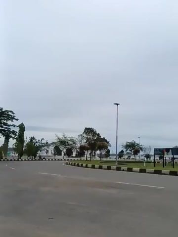 A video captured from a vehicle shows a paved road curving to the left, bordered by a median strip with black and white markings. Trees are planted along the median. In the background, there are some buildings and more trees. A lamppost stands tall in the middle distance. Two motorcycles pass by. The first motorcycle, ridden by a single person, moves from left to right across the frame. The second motorcycle, carrying two people, also travels from left to right, slightly further away. The sky is overcast, with gray clouds filling the frame.