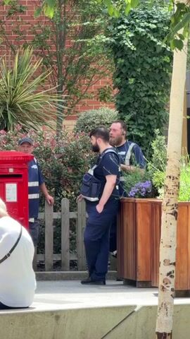 The video opens with a view of two men in dark blue uniforms, one with a backpack, standing near a wooden planter filled with purple flowers. Behind them, a fence separates them from a red post box and lush greenery. A man in a white sleeveless shirt is visible further back, facing away from the camera, with a woman in a white t-shirt sitting in the foreground. The scene appears to be outdoors, possibly a public area with shops or businesses, indicated by the brick building and modern wooden planters. The lighting suggests a bright, sunny day. As the video progresses, the camera pans slightly, revealing a woman in a yellow dress and sunglasses talking to the woman in the white t-shirt. Another man in a blue uniform is partially visible on the left side of the frame near the post box. The overall atmosphere is casual and relaxed, with people engaged in conversation or standing around outdoors.