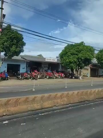 The video shows a roadside view of a business that sells tractors. Several red tractors are parked in front of the building. A motorcycle with two people on it is seen passing by. A dark colored car and a blue truck also drive past the scene. The weather appears to be clear with some clouds in the sky. A barrier with orange and white stripes is visible in the foreground.