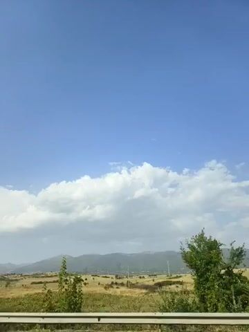 The video is a time-lapse recording of a wide landscape under a blue sky with some clouds. The foreground features a road barrier and dry vegetation. In the mid-ground, there are fields, scattered trees, and a small building. The background consists of rolling hills or mountains. The sun is visible, casting light on the scene. The camera appears to be stationary, capturing the slow passage of time.