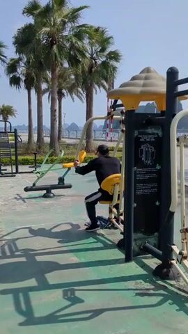 Here's a concise description of the video: 

A man is doing chest presses on a yellow and metal outdoor gym machine in what appears to be a park or public area. He's wearing a black jacket and pants with a white stripe, and a black baseball cap. Behind him are several palm trees and a view of water with rock formations in the distance, indicating a seaside or coastal location. A red and white striped pole stands further in the distance. Other pieces of exercise equipment are visible in the background. The area around the equipment is paved with a green and gray patterned surface. The sky is clear and blue.