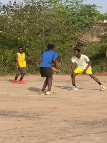 The video shows a group of young men playing basketball on a dirt court outdoors. Four players can be seen interacting around the basket. One player in a blue shirt is dribbling the ball, another in a white shirt is guarding him. A player in a yellow Lakers jersey is standing by. A fourth player wearing black shorts and a grey shirt jumps up in an attempt to block a shot. Other players can be seen in the background. The basketball hoop is rusty. Green trees and power lines can be seen surrounding the court.