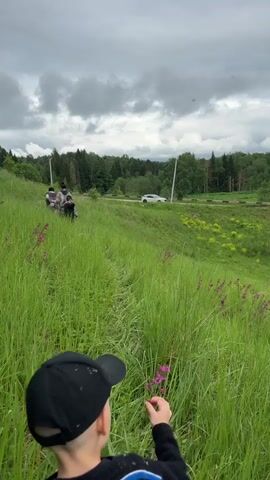 The back of a child wearing a black baseball cap and a striped sweater is visible in the foreground. The child briefly touches a pink flower with their right hand, then looks up towards a grassy hill. Two individuals, followed by another person, are walking away from the camera along a narrow path ascending the hill. The environment consists of tall green grass with scattered pink flowers, a distant road with a white car, power poles, and a treeline under a cloudy sky.