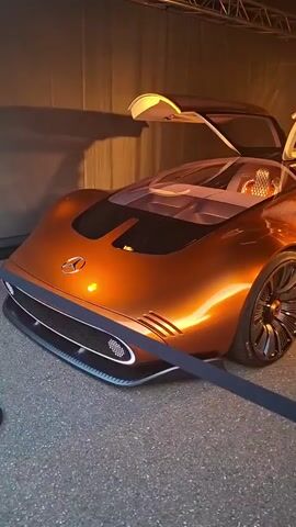 The video shows the exterior of a futuristic, orange Mercedes-Benz concept car with its gull-wing doors open. The car is parked on a paved surface, possibly a showroom or display area, with a dark backdrop. The camera pans slowly across the car, highlighting its sleek design, unique wheels, and illuminated interior. The lighting is dim, with spotlights illuminating the car.