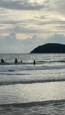The video shows a serene beach scene. Gentle waves roll onto the shore under a cloudy sky. In the water, several people are visible, wading and swimming. In the distance, a dark, tree-covered island or peninsula juts out into the sea. A small white object, possibly a boat or a parasail, can be seen near the island. The overall atmosphere is calm and tranquil, with the subdued light suggesting either early morning or late afternoon.