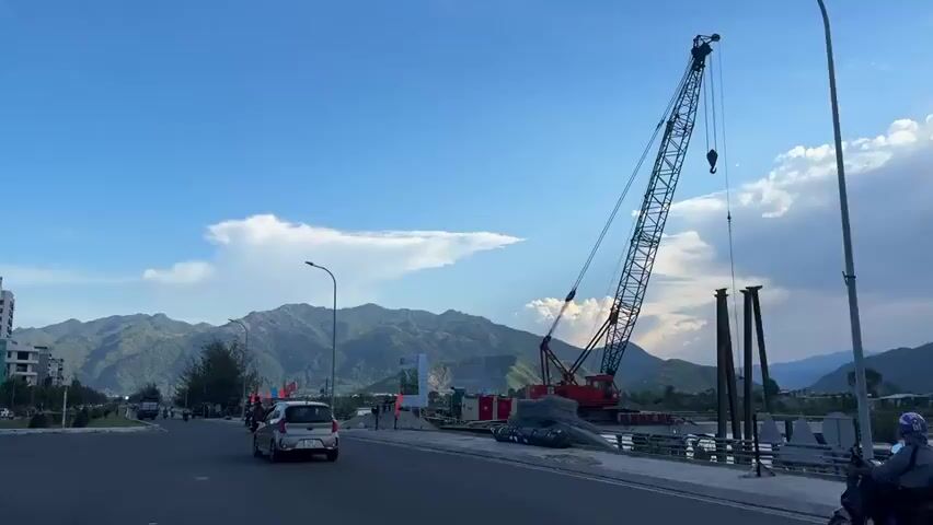The media file captures a scenic view of a coastal road with mountains in the background. The scene appears to be set in Nam Nha Trang Ward, Vietnam. In the foreground, a paved road is visible with several vehicles, including cars and motorcycles. The time of day is likely late afternoon or early evening, as suggested by the lighting and the quality of the sky, which is a vibrant blue with scattered clouds.

Dominating the right side of the frame is a large red construction crane, indicating ongoing development or infrastructure work, possibly a bridge or building project. Steel rebar and other construction materials are piled near the crane. Several tall, dark poles are also standing nearby, possibly part of the construction.

The middle ground features a slightly elevated embankment along the road, lined with trees and some temporary structures, perhaps market stalls or shelters. Colorful flags, some appearing to be national flags, are displayed. A few individuals are seen on foot and on motorcycles in this area, suggesting a moderately busy atmosphere.

In the distance, majestic, tree-covered mountains rise, creating a dramatic backdrop. A distinctive, large white cloud formation is prominent in the sky above the mountains. The overall impression is one of a developing area situated in a beautiful natural landscape. The presence of construction and traffic suggests normal daily activity. No discernible text is clearly visible within the frame.