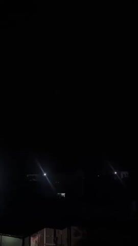 The video begins with a completely black screen. Lights appear in the distance, casting beams. The scene then transitions to a dark sky with visible clouds and purple hues, suggesting a storm. Distant lights are still visible in the silhouette of buildings and structures. The video returns to a black screen with lights in the distance.