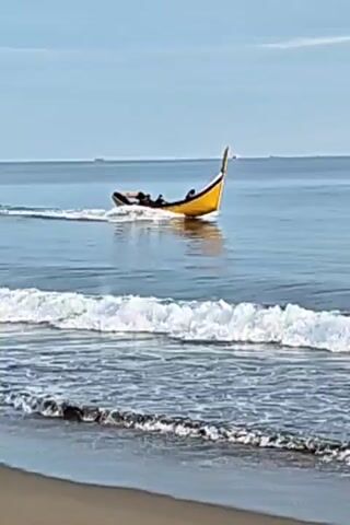 A small yellow boat with a tall fin is moving rapidly across the water, leaving a wake behind it. The boat is occupied by at least three people who appear to be sitting down. In the foreground, waves are crashing onto a sandy beach. The sky is a pale blue, and the water is a deep blue.