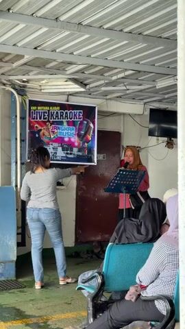 The video shows a woman in a hijab singing karaoke on a boat. She is standing behind a music stand with a microphone in hand. There is a banner behind her that says “Live Karaoke.” Another woman in jeans is standing near the banner and pointing to it. Several people are sitting in blue chairs, watching the karaoke performance. The boat has a low ceiling with metal beams. A television is mounted on the wall behind the singer. A dark door is to the side of the banner.
