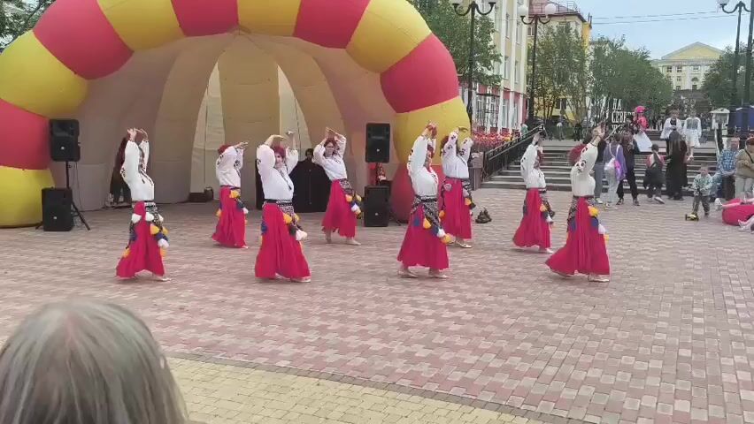 A group of nine women are performing a folk dance in an outdoor public space. They are dressed in traditional Ukrainian attire, consisting of white embroidered blouses, red skirts with decorative fringes, and red headbands. The women are arranged in a semi-circle formation in front of a large, inflatable archway that is yellow and red. Two speakers are positioned to the left of the dancers. The ground is paved with interlocking stones. In the background, there are buildings, trees, and other people observing the performance. Some onlookers are visible in the foreground, with the back of one person's head in focus. The lighting suggests it is daytime.