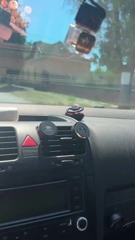 The video shows a close-up view of the interior of a car, focusing on the dashboard and windshield. The camera pans across the dashboard, revealing a car's air vent with two magnetic phone mounts attached. One mount has a circular black piece with the number 89 and the word AROMATIC. The other mount is a black circle with a gold accent in the shape of an A. To the right of these mounts, there is a spherical air freshener with red accents. Outside the car, a yellow house with a brown roof and green trees are visible through the windshield. The sky is blue and clear, suggesting a sunny day.
