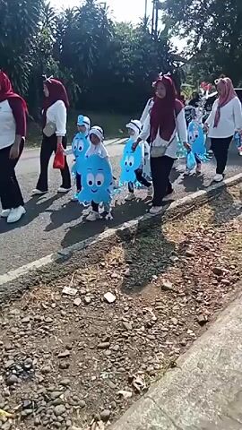 The video shows a group of people marching in a parade. The people in the front are children dressed in blue octopus costumes. They are accompanied by adult women wearing hijabs and white shirts. The parade is taking place on a paved road with a gravel shoulder. There are trees and foliage in the background, suggesting an outdoor setting. The lighting indicates it is daytime.