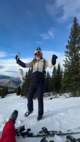 A woman in ski gear stands on a snow-covered slope, throwing a white object in the air, while another person, wearing red ski pants and ski boots, is lying down. The woman is wearing a white and black ski jacket, navy blue ski pants, a white helmet, and goggles. She is holding a phone in one hand. Behind her, there are evergreen trees and a view of mountains under a blue sky with scattered clouds.  Other skiers and snowboarders are visible in the distance.