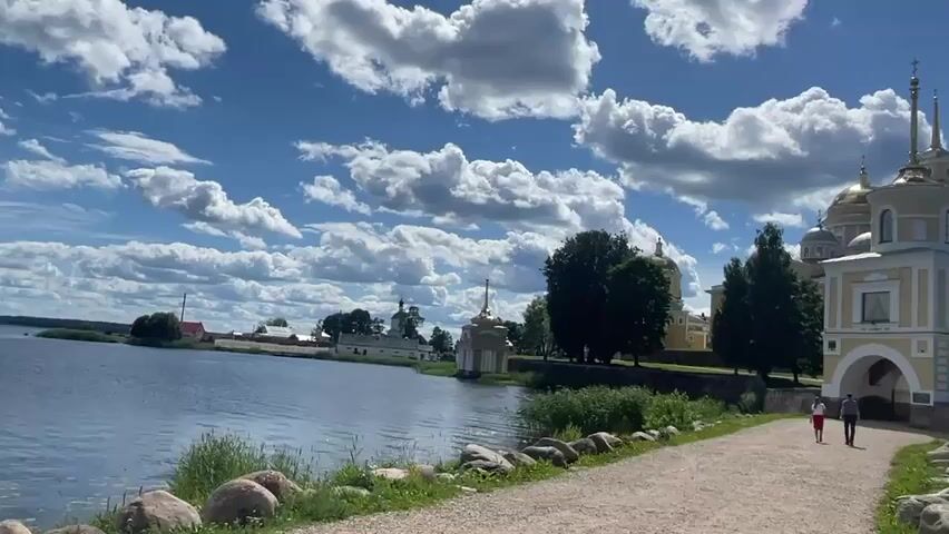 This is a video clip showing a scenic view of a lake with a monastery or church complex in the background. The sky is a vibrant blue with large, fluffy white clouds, suggesting a clear and pleasant day, likely mid-morning or afternoon due to the bright sunlight casting shadows. The sun is quite bright, creating a strong contrast and highlighting the architectural details of the buildings.

The foreground features a gravel path winding along the edge of the water, lined with large rocks and green grass. Two people are walking away from the camera on the path, one in a red top and white bottoms, the other in a dark top and lighter bottoms. They appear to be tourists or visitors enjoying a stroll.

The main subject is the impressive religious complex situated on the opposite side of the lake. It features several onion-domed buildings, characteristic of Russian Orthodox architecture, painted in yellow and white with golden spires that glint in the sunlight. A prominent gatehouse-like structure with an arched entrance stands closest to the water on the right side of the frame. Trees surround the buildings, adding to the picturesque setting.

In the distance, across the calm water, more buildings of the complex are visible, including a white church with a blue dome. The overall atmosphere is peaceful and serene, evoking a sense of tranquility and historical grandeur. The location cues, such as the architecture and the presence of a large body of water, combined with the provided context of Svetlitsa, Russia, confirm this as a likely view of a Russian monastery or church complex by a lake. There is no visible text in the video.