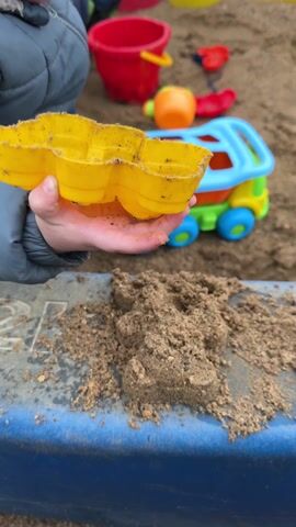 A child in a gray jacket plays in a sandbox, using a yellow plastic mold to shape the wet sand. The sand, mixed with small pebbles, is piled on a blue ledge. In the background, a colorful plastic toy truck sits in the sandbox, surrounded by a red bucket, a yellow spoon, and other assorted toys. The child places the yellow mold into the sand, then lifts it up.